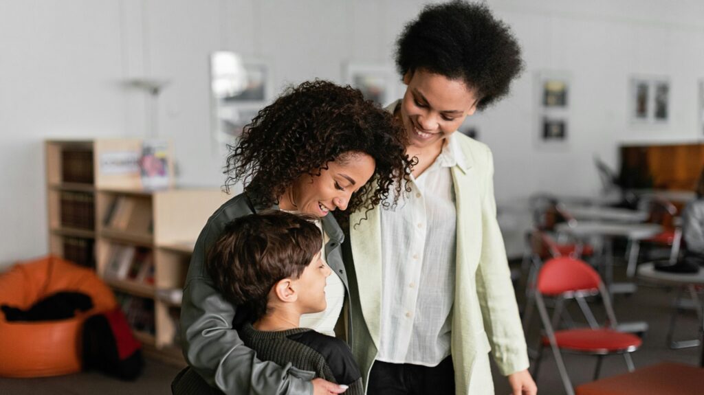 "Two women smiling and embracing a young boy in a school setting."