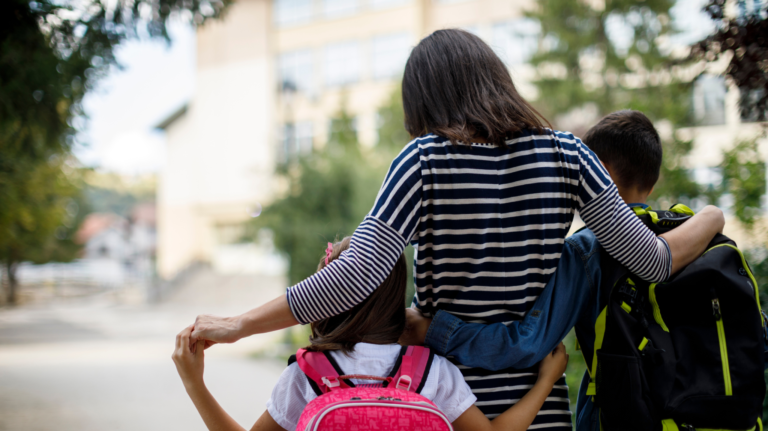 Woman stands with her arms around two children wearing school backpacks