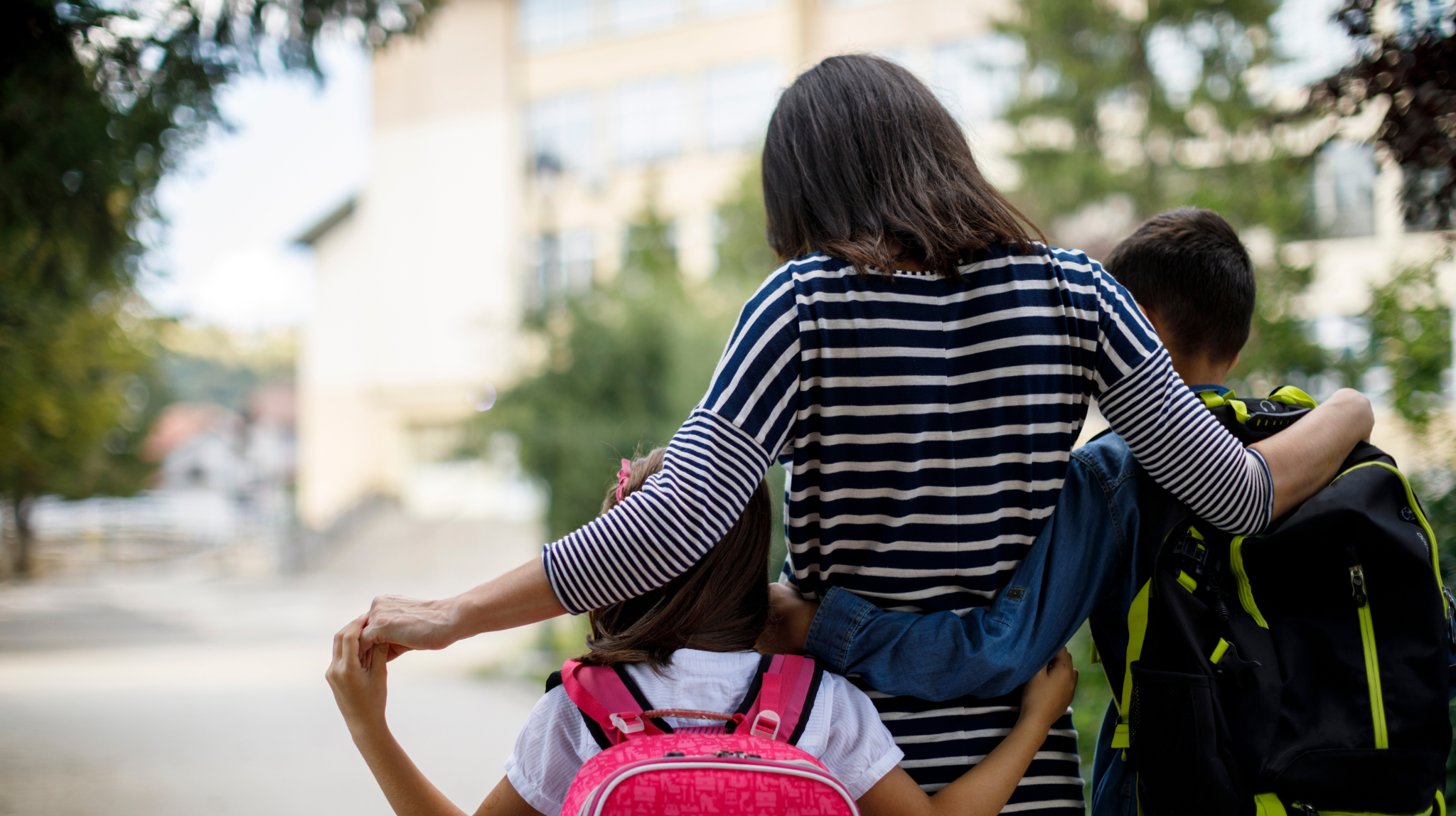 Woman stands with her arms around two children wearing school backpacks